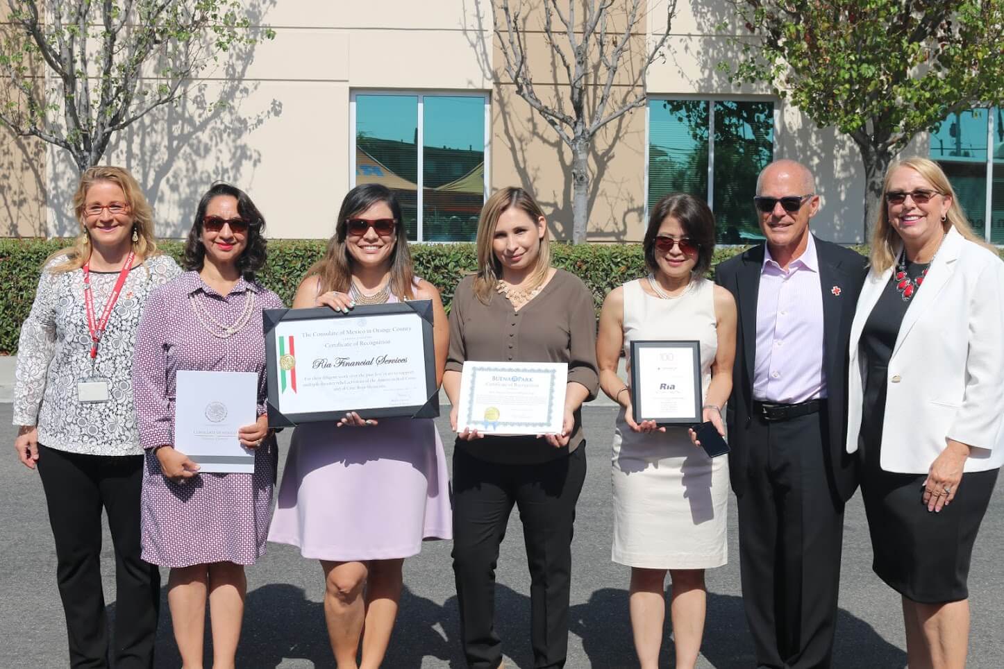 A group photo features seven smiling people standing outdoors in front of a building. Four individuals hold certificates and plaques, indicating awards or recognition—perhaps from a successful blood drive campaign. They are dressed in business casual attire, with trees and a clear sky visible in the background.
