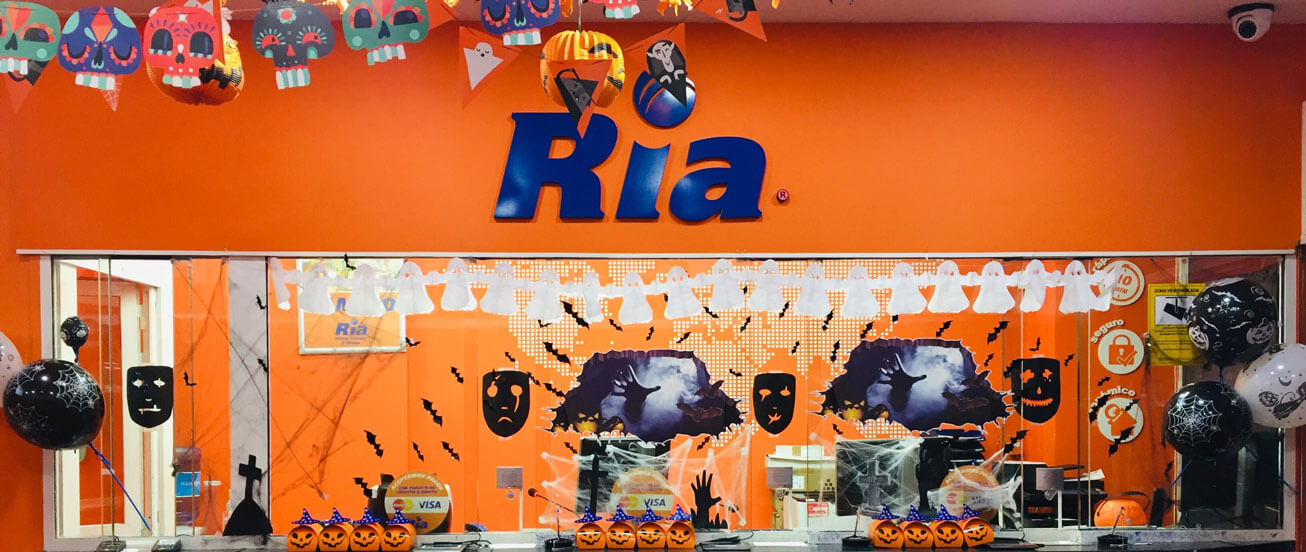 A room is decorated for Halloween with orange and black-themed decor, including masks, bats, and cobwebs. The wall has a large "Ria" sign, and the counter displays carved pumpkin faces. Banners and hanging decorations add to the festive atmosphere.
