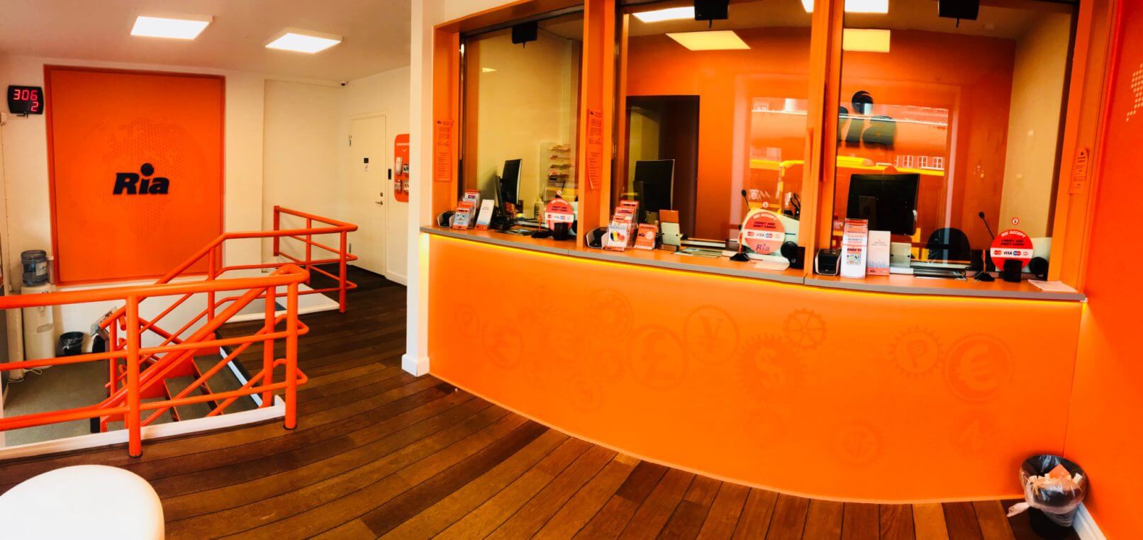 A brightly lit office with orange walls and wooden floors showcases Ria’s Formula for Employee Management. The office features a counter with several windows and a staircase with orange railings leading downstairs. Various currency symbols are displayed on the orange walls, and "Ria" is printed on the left wall.