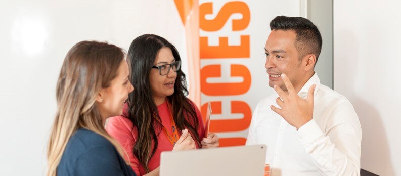 Three people engaged in a lively discussion stand near a whiteboard with an orange banner displaying part of the word "SUCCESS." Two women, one in a pink shirt and the other in a blue top, listen attentively as a man in a white shirt explains Agile scrum methodology while gesturing with his hand.
