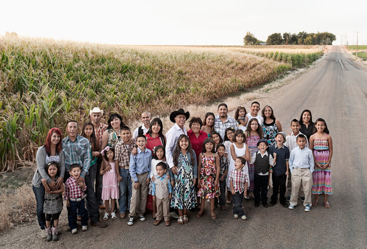 A large multi-generational family stands on a road next to a cornfield. The group includes children, teens, adults, and elderly members, all smiling and dressed in casual, colorful clothing. The sky is clear, and the scene is set during daylight hours.