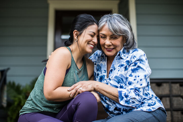 A middle-aged woman and an older woman sit closely together on a porch, sharing a warm embrace and smiling. The middle-aged woman wears a green sleeveless top, while the older woman sports a patterned blouse. This heartfelt moment could inspire Mother's Day ideas from far away, capturing the essence of connection despite distance.