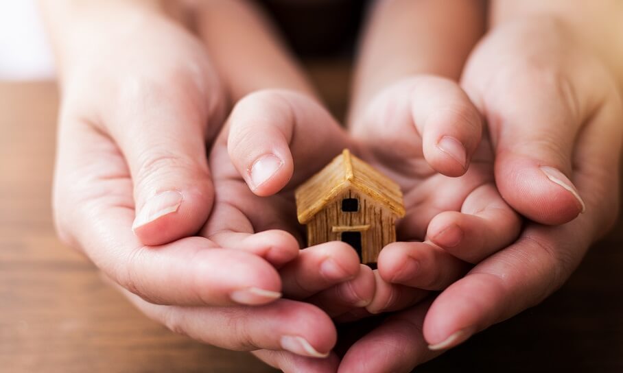Close-up of two pairs of hands cupping a small wooden house. The larger hands appear to be those of an adult, while the smaller ones seem to be those of a child. The focus is on the tiny house model, symbolizing care, protection, or family.
