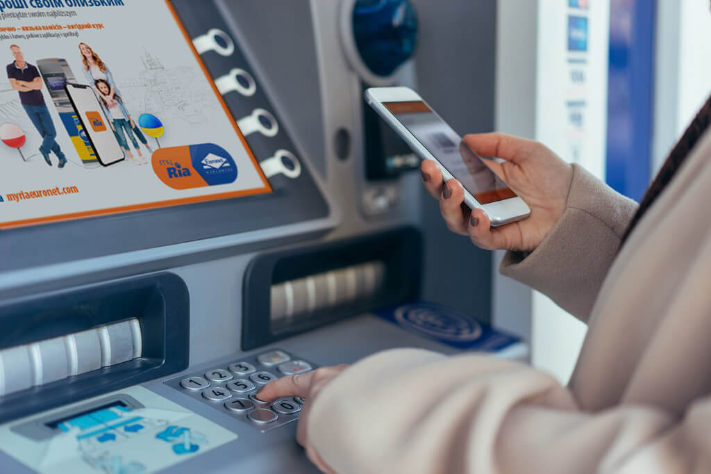 A person stands at an ATM, pressing buttons on the keypad with one hand while holding a smartphone with the other. The screen on the ATM displays transaction options, and an advertisement is partially visible on the left side of the machine.