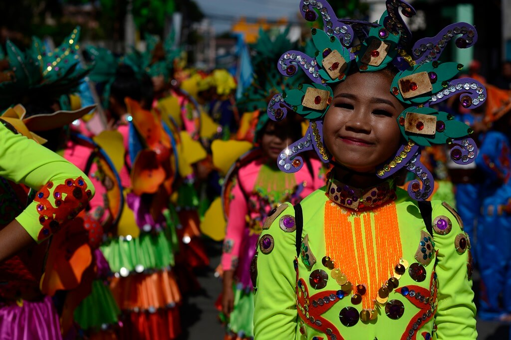 A person dressed in a vibrant, colorful costume adorned with sequins, beads, and flowers smiles while participating in a lively street parade. Members of various hometown associations join them in the background, creating a festive and cheerful atmosphere.