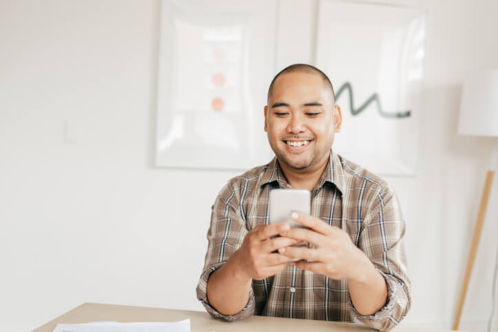A smiling man sits at a table, holding his smartphone. He is wearing a plaid shirt and is in a well-lit room with minimalist decor, including framed artwork and a floor lamp. He looks pleased, perhaps ready to send money to the Philippines effortlessly through his phone.