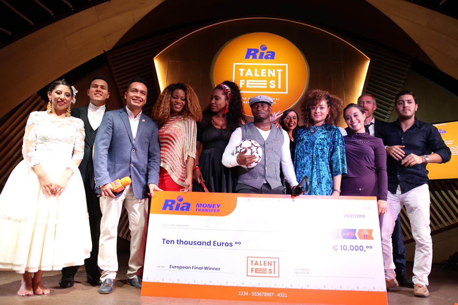 A group of people pose on stage around a large ceremonial check for ten thousand Euros, with a "Ria Talent Fest" banner in the background. Some of them hold microphones and a soccer ball, appearing to be smiling and celebrating their success at the Ria Talent Fest.