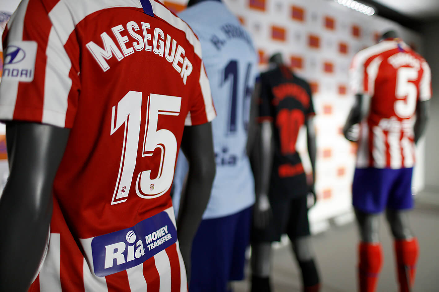 A lineup of mannequins displaying different soccer jerseys, with the focus on a red and white striped Atlético de Madrid jersey featuring the name "Meseguer" and number 15. Other jerseys with varying colors and numbers are also visible in the background.