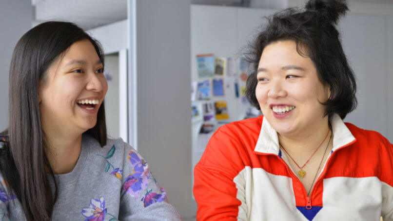 Two women in science are sitting and laughing together indoors. The woman on the left wears a grey top with floral print, and the woman on the right wears a red and white jacket. In the background, there are blurred images and notes pinned to a surface.