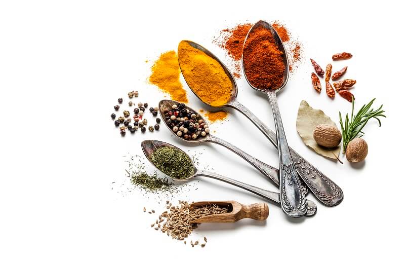 A variety of spices displayed on spoons and scattered around on a white surface, evoking the essence of Indian food. The spices include vibrant turmeric, paprika, peppercorns, dried herbs, and small red chili peppers, with a nutmeg, bay leaf, and rosemary sprig alongside.