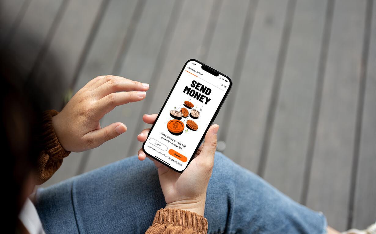 A person holding a smartphone on their lap uses the Ria app money transfer feature, with the text "Send Money" and an orange send button displayed on the screen.