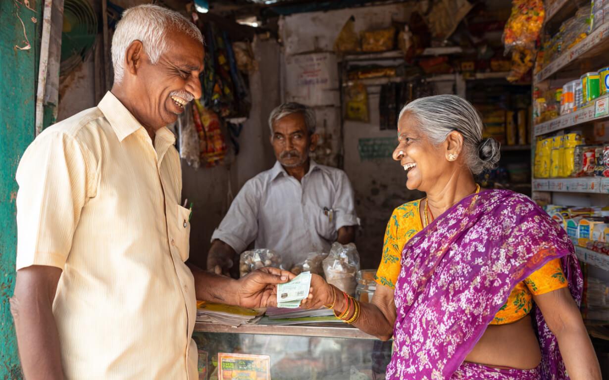 An elderly man and woman smile as she hands him a card inside a small, colorful shop with shelves of goods. A shopkeeper stands behind the counter, observing their Send Money to India transaction.
