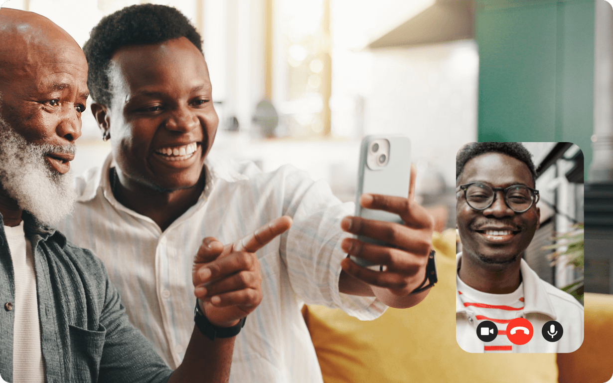 Dos hombres sonríen y señalan un smartphone mientras chatean por vídeo con otro hombre sonriente, visible en una pequeña pantalla superpuesta. Parecen felices y están en el interior, sentados en un sofá amarillo.