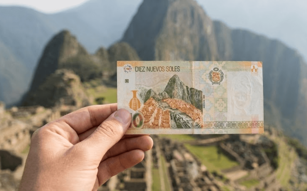 A hand holds Peruvian currency—a 10 soles banknote—in front of Machu Picchu, aligning the image on the note perfectly with the actual ancient Inca ruins and majestic mountains in the background.