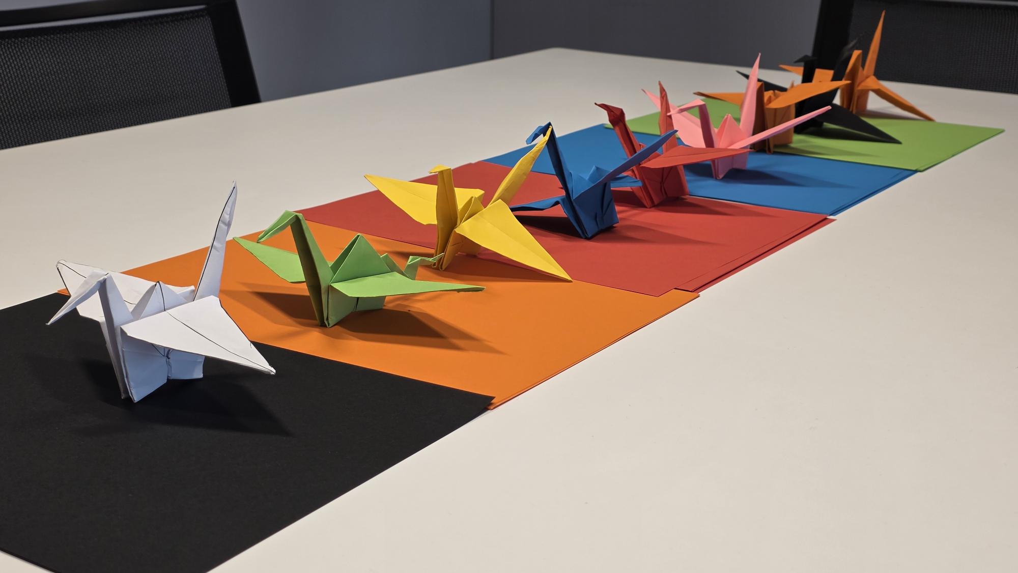 Several colorful origami cranes are arranged in a line on sheets of colored paper, forming a rainbow spectrum, on a white tabletop with black chairs in the background.